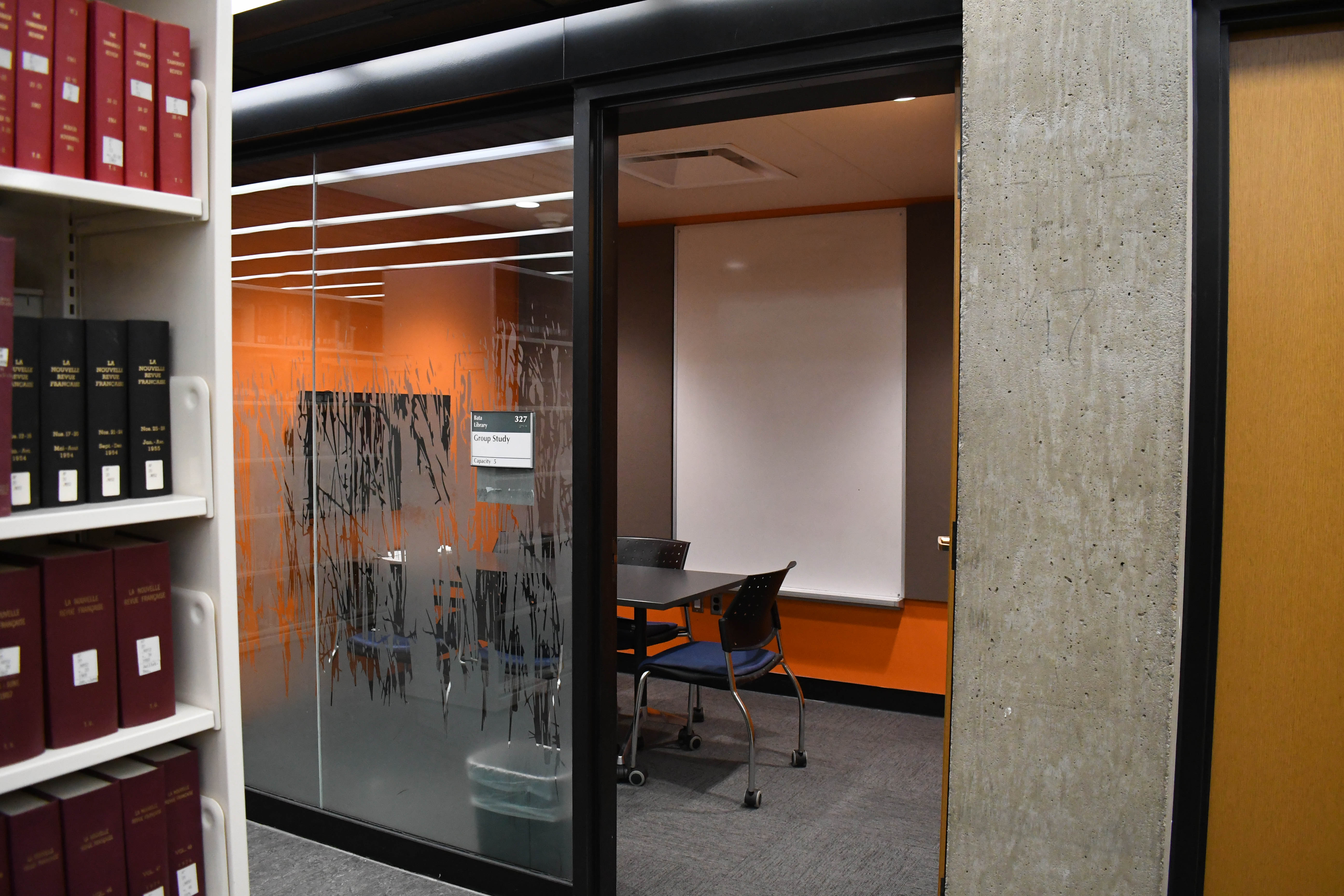 A modern library study space with orange walls, featuring a table and chair set against a backdrop of bookshelves filled with volumes and a glass door marked 'Group Study'.