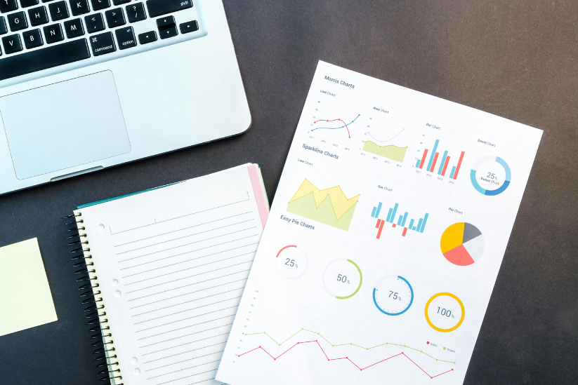 An open notebook beside a laptop with colorful charts and graphs printed on paper, indicating data analysis.