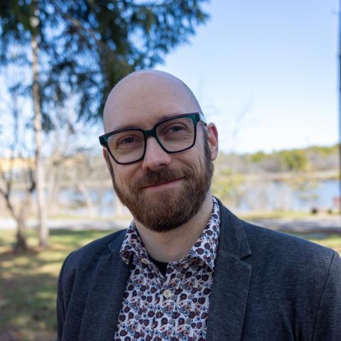 A man with a beard and glasses, dressed in a patterned shirt and dark blazer, stands outdoors with a background of trees.