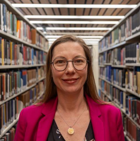 A woman with long straight hair and glasses, wearing a bright pink blazer over a floral top, smiles in a library.