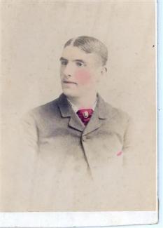 Photograph of a man posing. Photograph is mostly in black and white but he has pink cheeks, a red scarf and pocket square