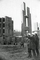 Three people in construction helmets pointing at a building, with someone walking by in the background